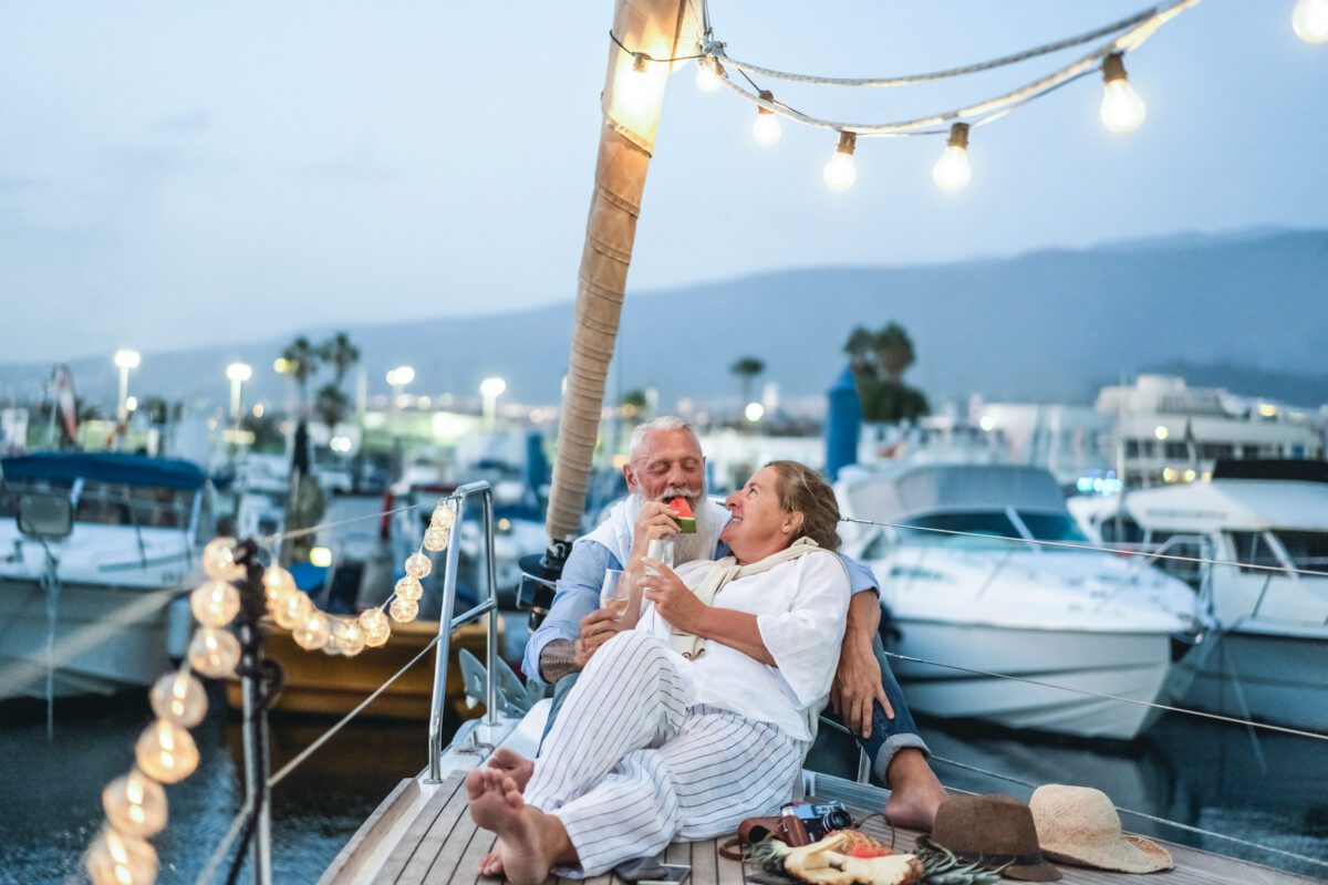 BRAIN Yachting Entschleunigen Senior couple drinking champagne and eating fruits on sailboat during summer vacation - Focus on faces